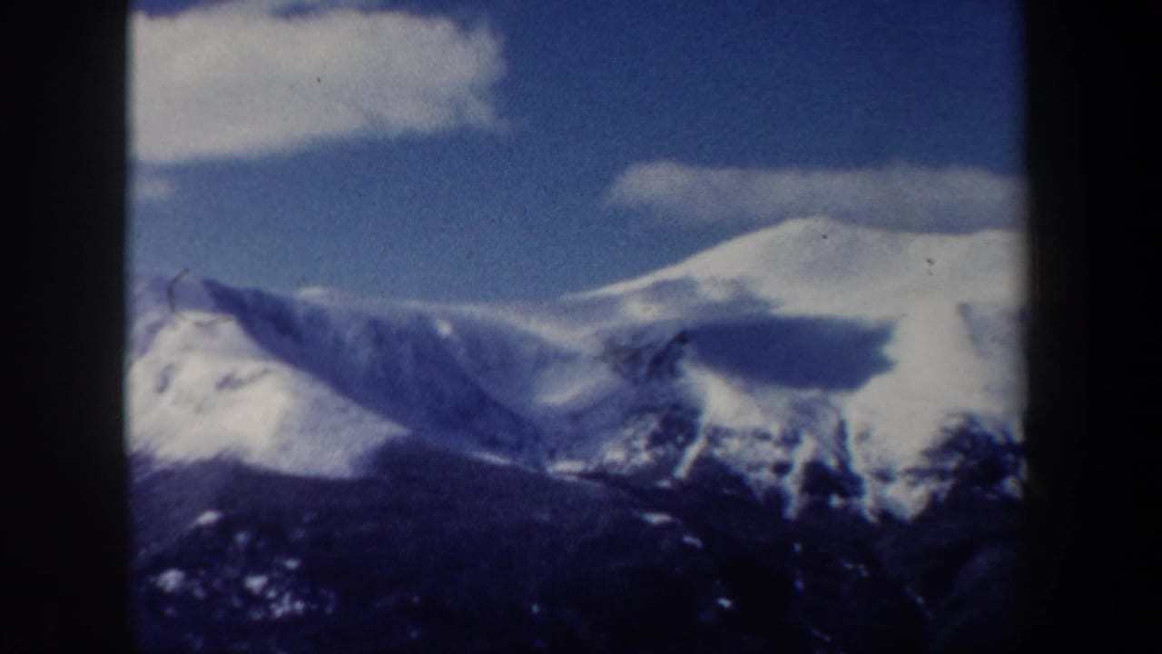 1962 New Hampshire 8mm frame showing a wider snow-covered mountain scene, dark foreground trees, and clouds over the ridge.