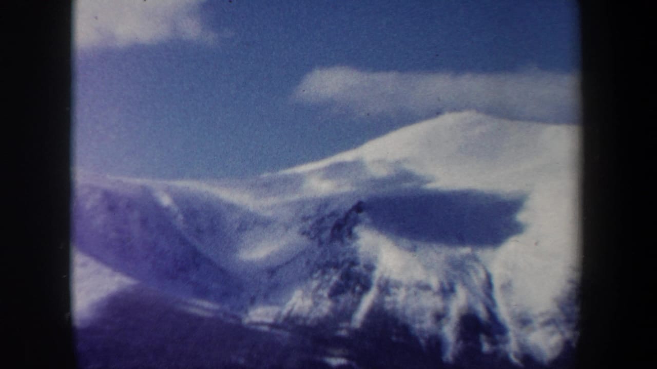 1962 New Hampshire 8mm frame of a rounded snowy peak, cloud bands, blue sky, and dark forested foreground.