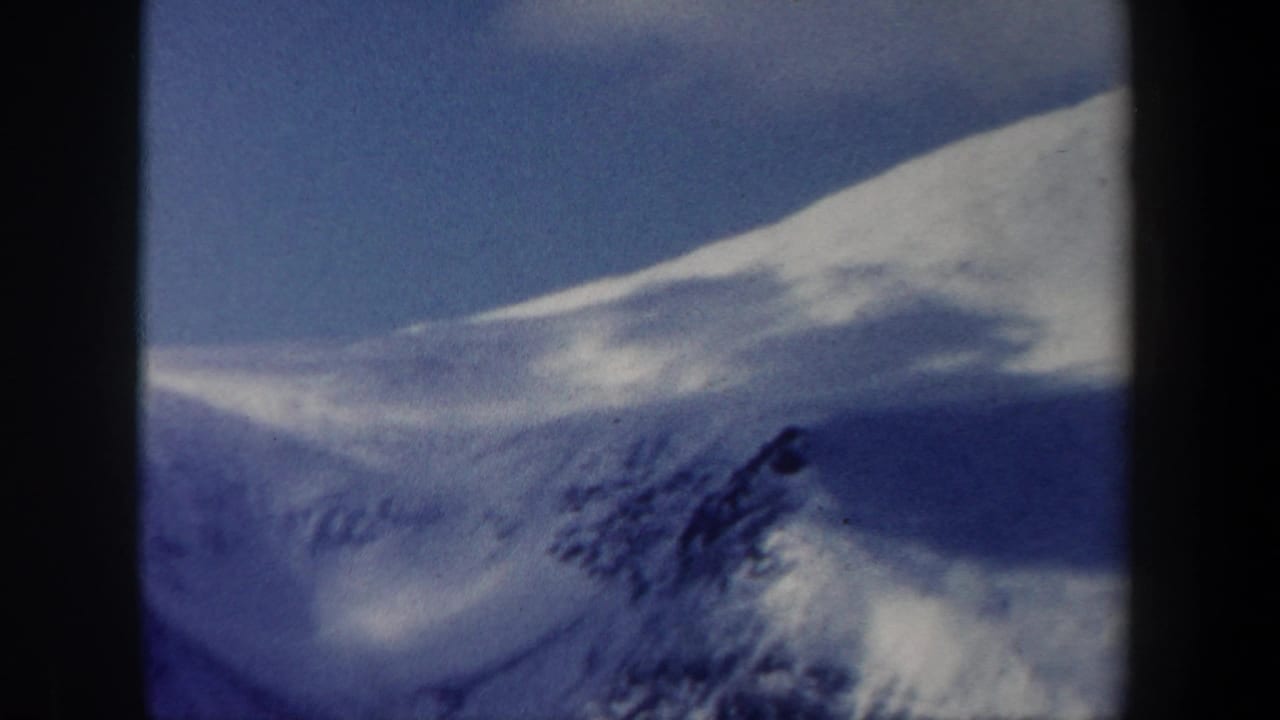 1962 New Hampshire 8mm frame of a snow-covered mountain slope, dark outcrop, and clear blue winter sky.