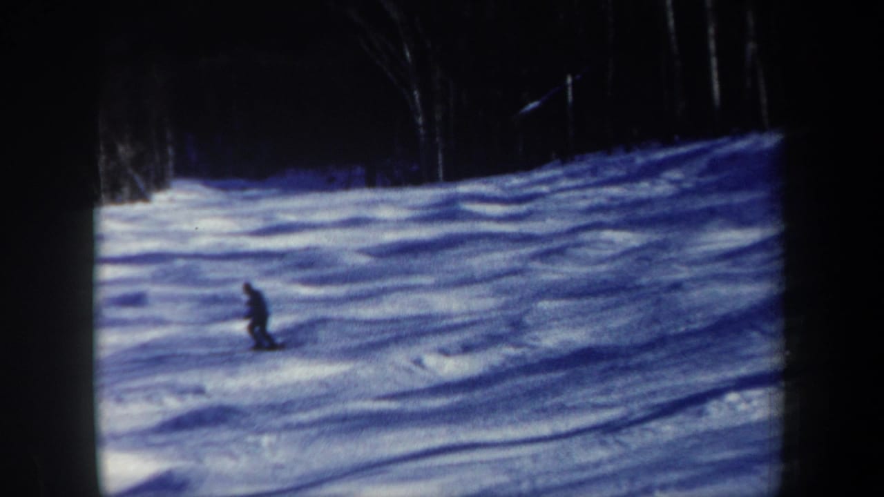 The skier moves steadily down the snow-covered slope, framed by dark trees and 8mm projector borders.