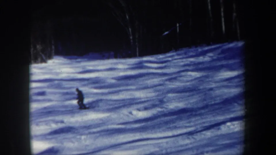 The skier moves steadily down the snow-covered slope, framed by dark trees and 8mm projector borders.