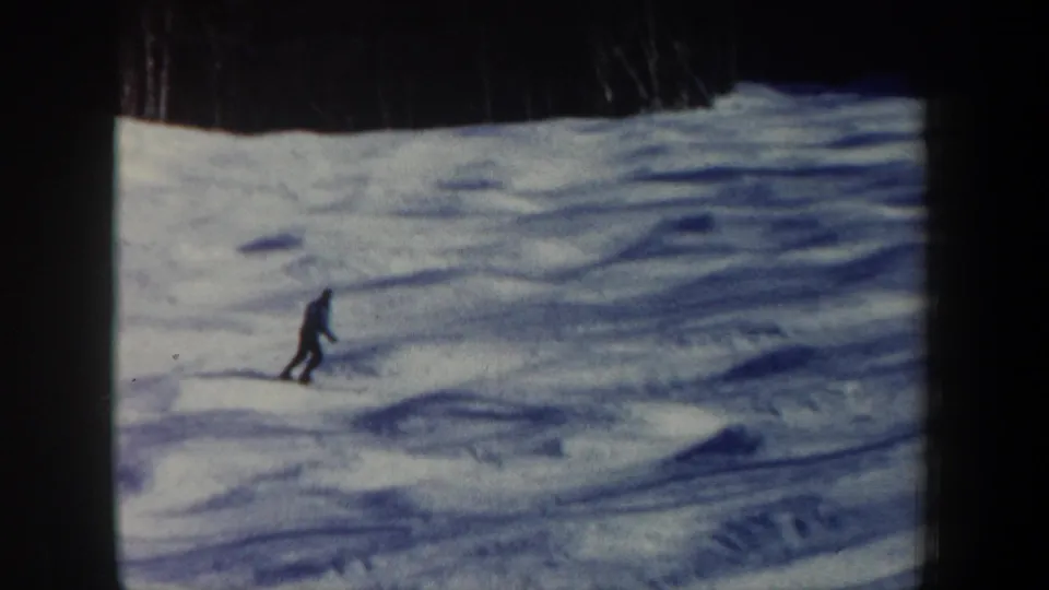 The skier reaches a lower part of the snowy hillside, still distant within the dark 8mm frame mask.