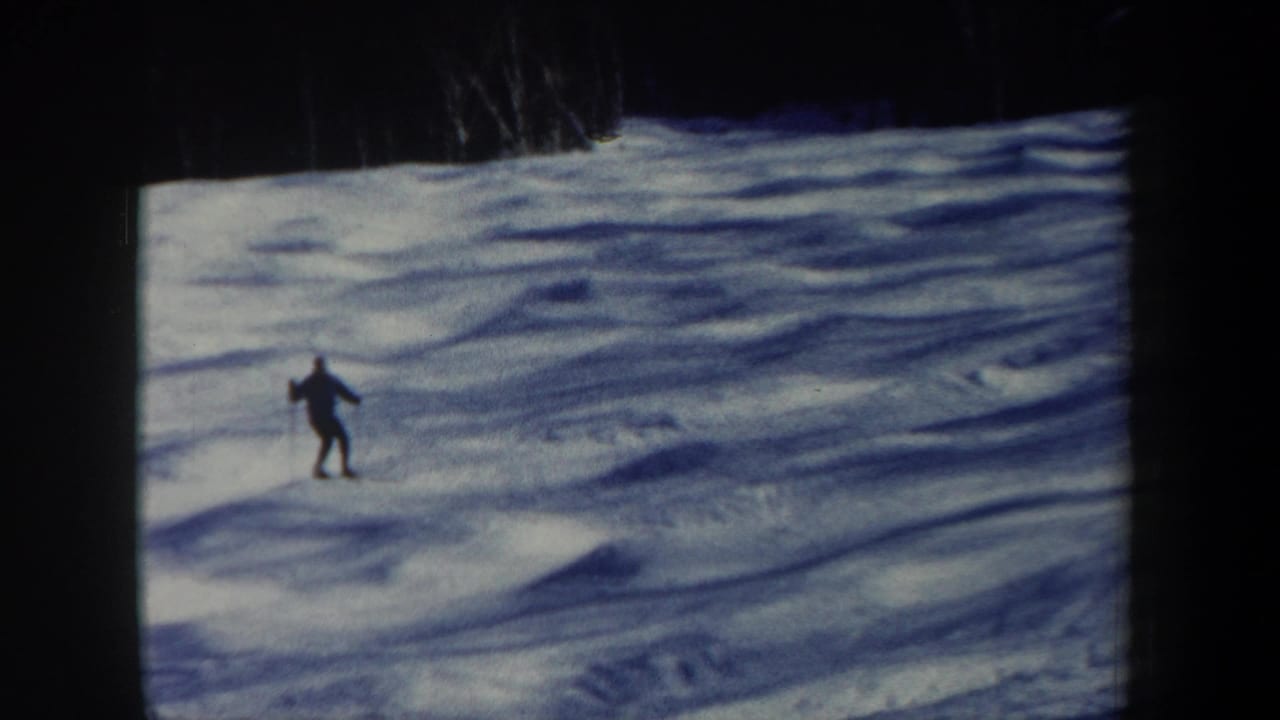 A distant skier descends a snowy New Hampshire slope in 1962 8mm footage, with blue shadows and a dark treeline above.