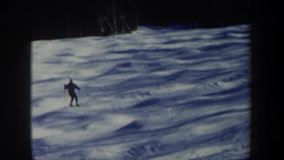 A distant skier descends a snowy New Hampshire slope in 1962 8mm footage, with blue shadows and a dark treeline above.