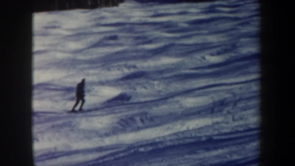 A distant skier continues across the lower snowy slope in vintage 8mm footage from New Hampshire, with dark clothing against pale snow.