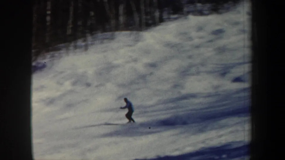 1962 New Hampshire 8mm frame showing the lone skier farther downhill, filmed from a distant home-movie viewpoint on a wooded snow slope.