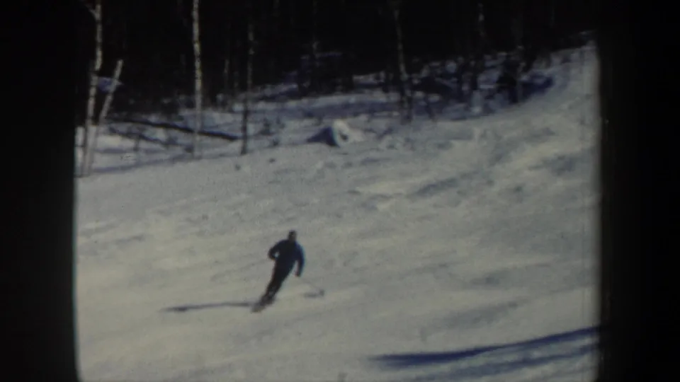 1962 New Hampshire 8mm frame of a lone skier carving across the lower right of a broad snowy slope below a dark stand of trees.