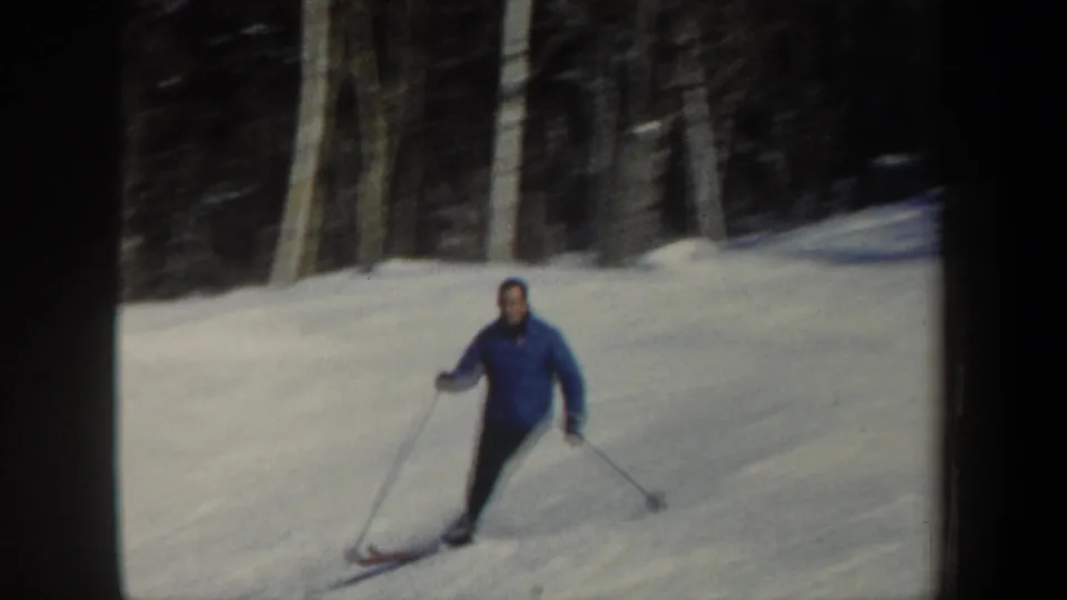 The skier approaches the camera on a snowy wooded slope, wearing a blue jacket and dark pants.