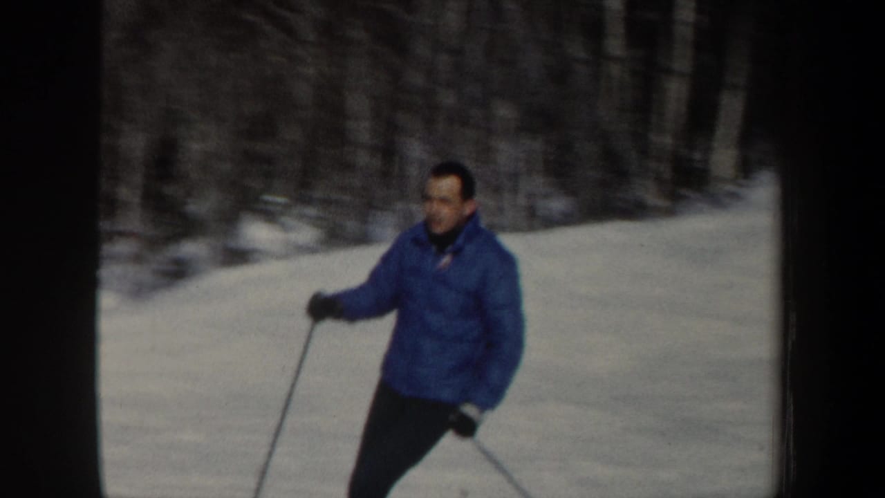 Closer 1962 New Hampshire frame of a man in a blue winter jacket skiing past a wooded snowbank.