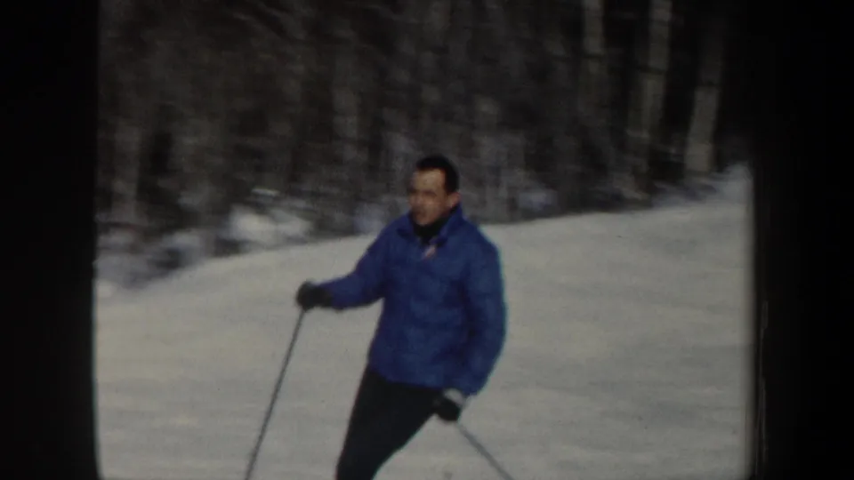 Closer 1962 New Hampshire frame of a man in a blue winter jacket skiing past a wooded snowbank.