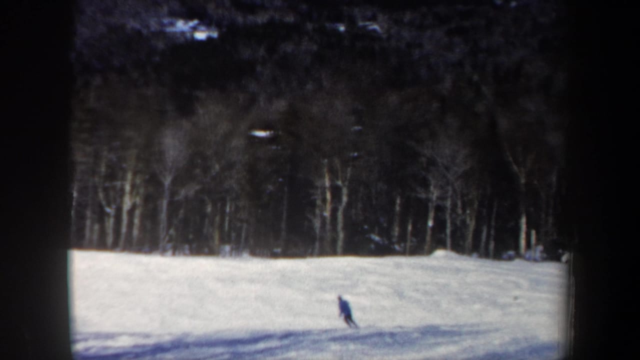 A skier in blue continues across the lower slope, framed by snow, shadows, and bare New Hampshire woods.