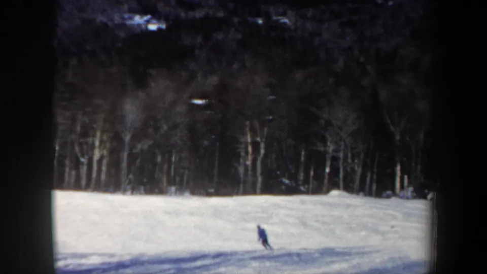 A skier in blue continues across the lower slope, framed by snow, shadows, and bare New Hampshire woods.
