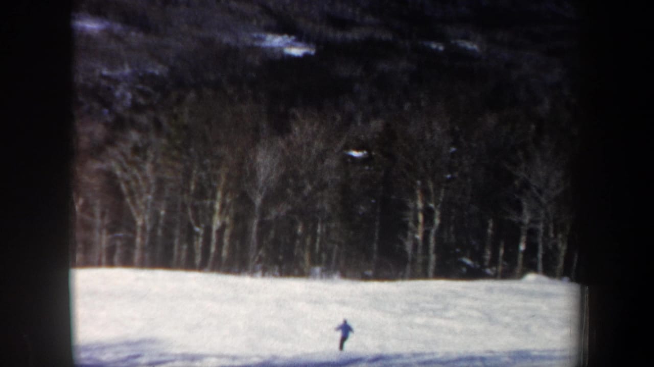1962 New Hampshire home-movie frame showing a distant skier in blue on a white slope bordered by bare trees.