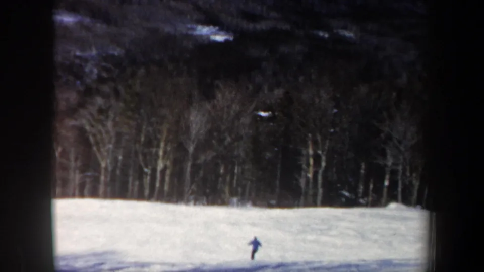 1962 New Hampshire home-movie frame showing a distant skier in blue on a white slope bordered by bare trees.