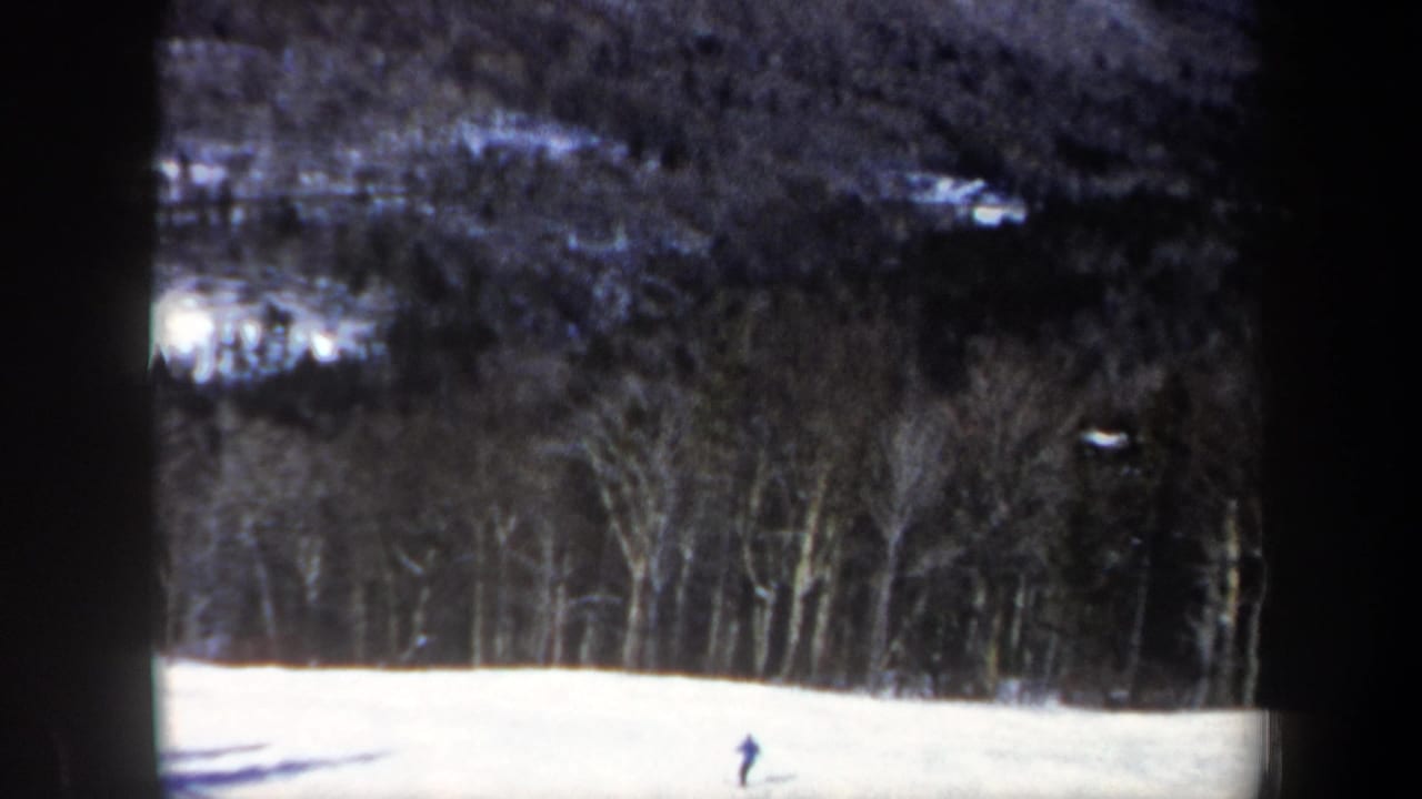 Archival 8mm frame from New Hampshire showing a lone skier near the lower part of a snowy hill, backed by winter woods.