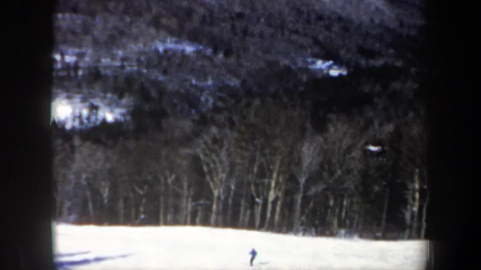 Archival 8mm frame from New Hampshire showing a lone skier near the lower part of a snowy hill, backed by winter woods.