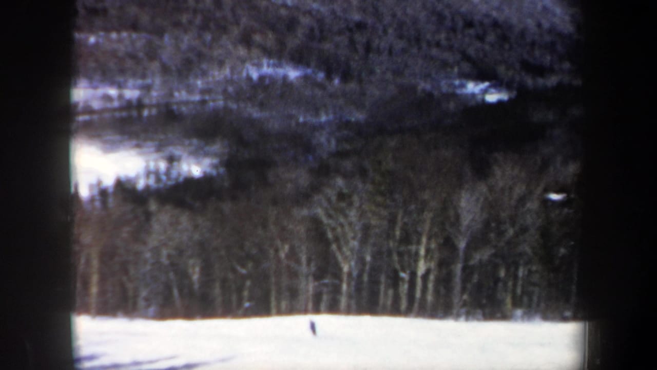 1962 New Hampshire home-movie frame showing a distant skier on a snowy slope beneath bare trees and forested hills.