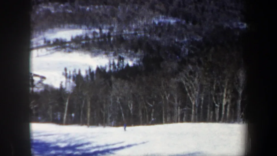 A small skier moves across a New Hampshire ski slope, with bare trees and dark wooded hills behind the snow.