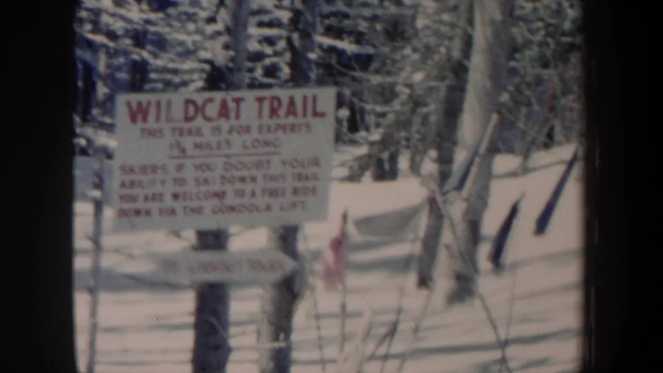 1962 New Hampshire 8mm still of a Wildcat Trail warning sign amid snow, bare trees, ski poles, and a small red flag.