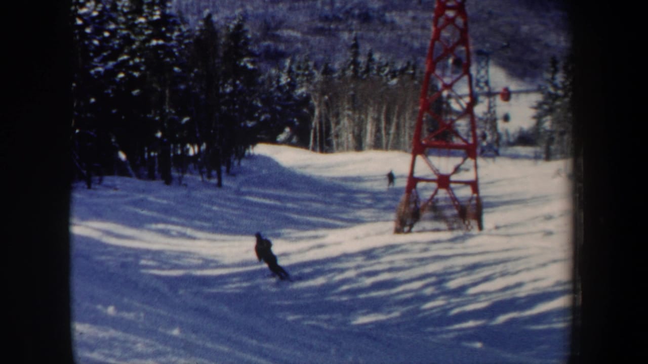 A skier cuts across the snowy slope beneath a red lift tower, with evergreens and winter light in the background.