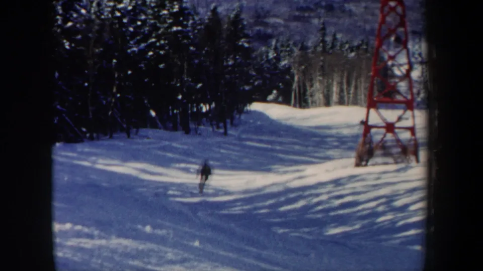1962 New Hampshire 8mm frame showing a distant skier crossing a sunlit snowy slope beside evergreens and a red ski lift tower.