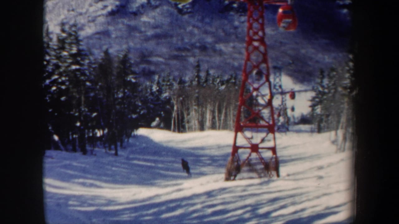 1962 New Hampshire 8mm view of a skier farther down the slope, with blue-shadowed snow, trees, and a red lift tower in frame.