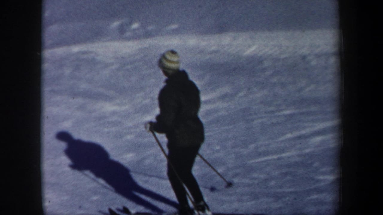 Closer 1962 New Hampshire frame of a skier in dark clothing and a light striped knit hat, standing on skis with poles and a long shadow.