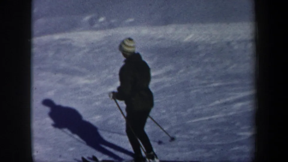 Closer 1962 New Hampshire frame of a skier in dark clothing and a light striped knit hat, standing on skis with poles and a long shadow.