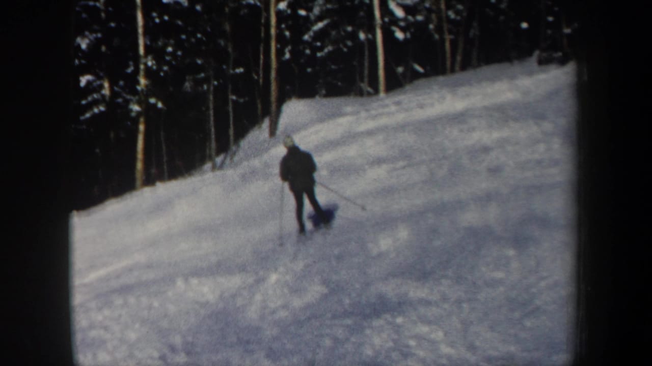 A distant skier continues across a wide snowy slope, framed by tall trees and soft handheld 8mm focus.