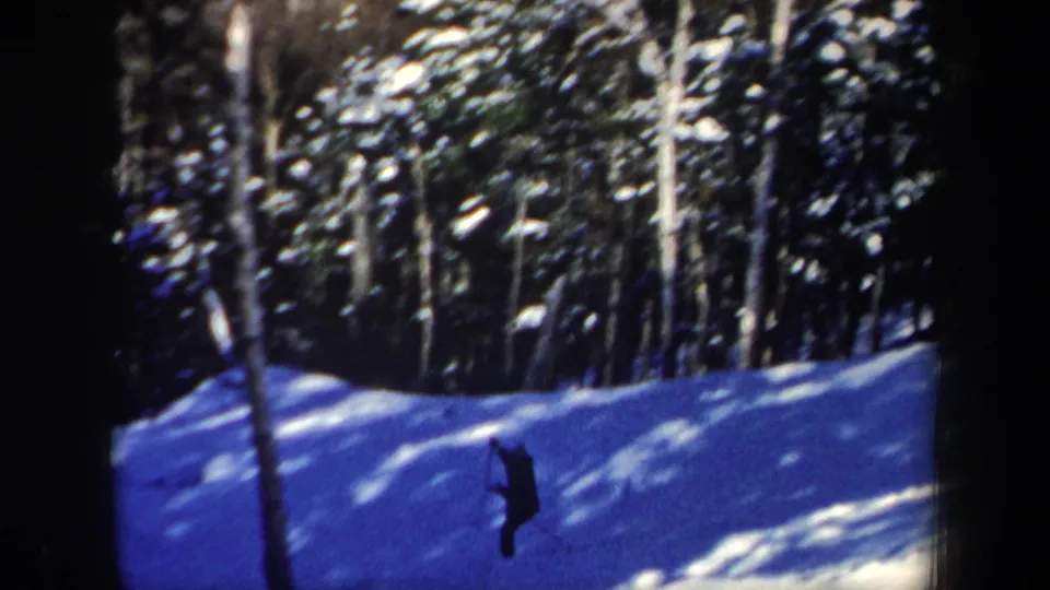 A dark-clothed skier descends through trees on a bright snowy New Hampshire hillside in 1962 home-movie footage.