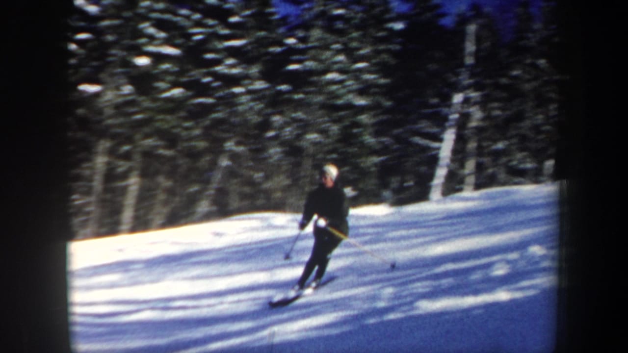 1962 New Hampshire 8mm frame showing a woman in dark winter clothing skiing across a sunlit snowy slope with poles.