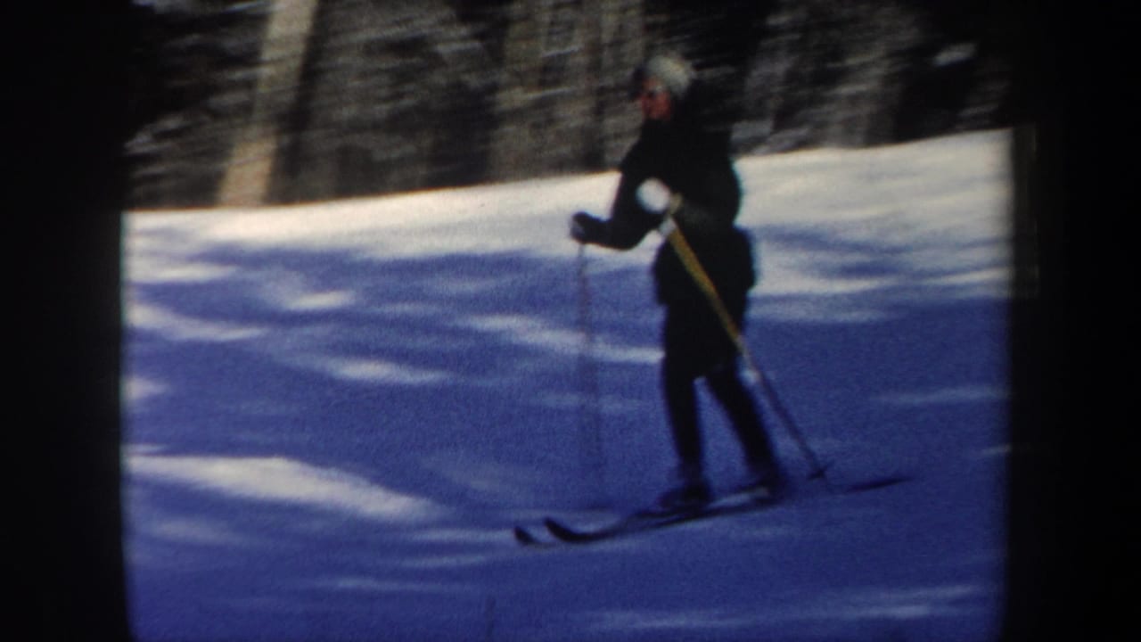 1962 New Hampshire home-movie frame with a closer view of a woman skier in a dark outfit and light hat on a wooded snowy slope.