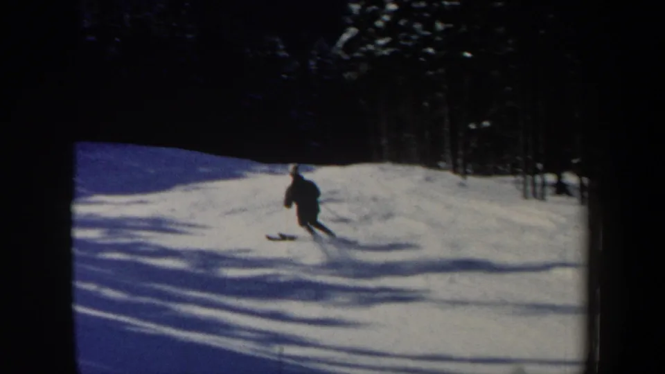 A small figure on skis continues downhill across bright snow, framed by dark trees and blue-toned shadows.