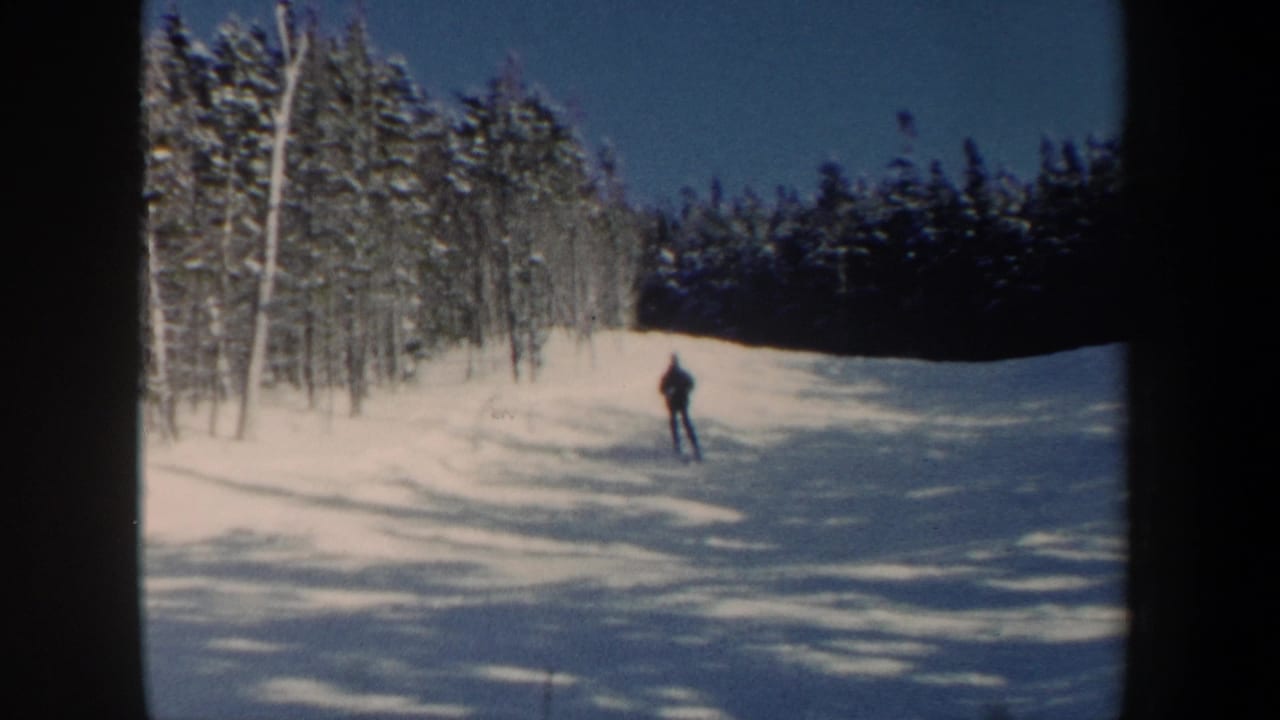 A lone skier in dark winter clothing descends a snowy New Hampshire hill, framed by trees and soft 8mm grain.