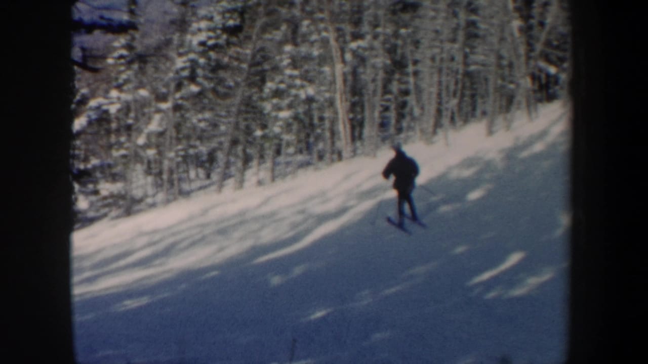 1962 New Hampshire frame with a distant skier descending through snow and long tree shadows in a forest setting.
