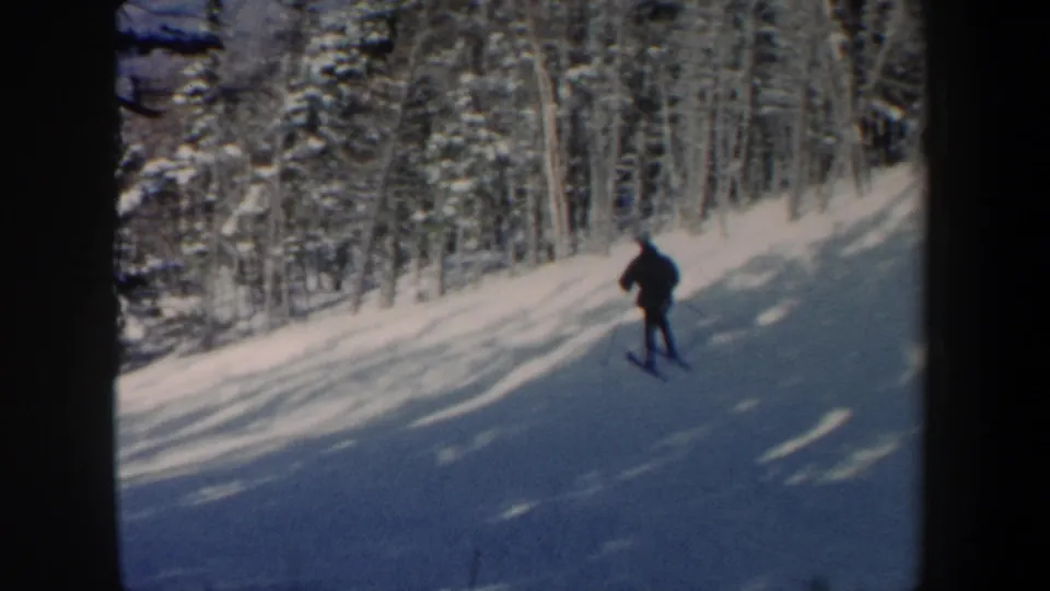 1962 New Hampshire frame with a distant skier descending through snow and long tree shadows in a forest setting.