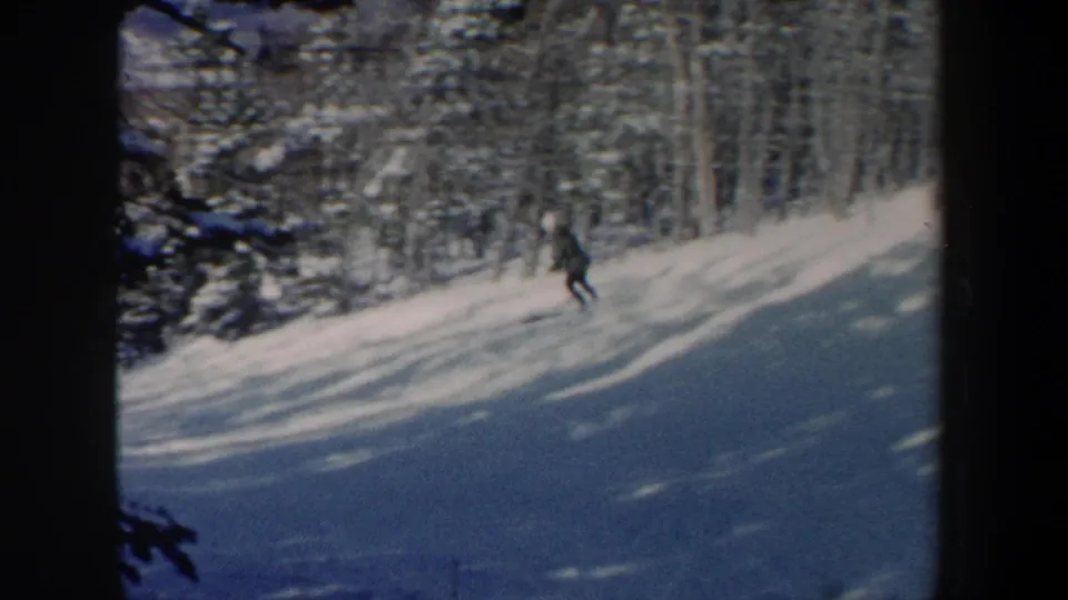 1962 New Hampshire 8mm frame showing a distant skier moving downhill across a snow-covered slope near dense evergreens.