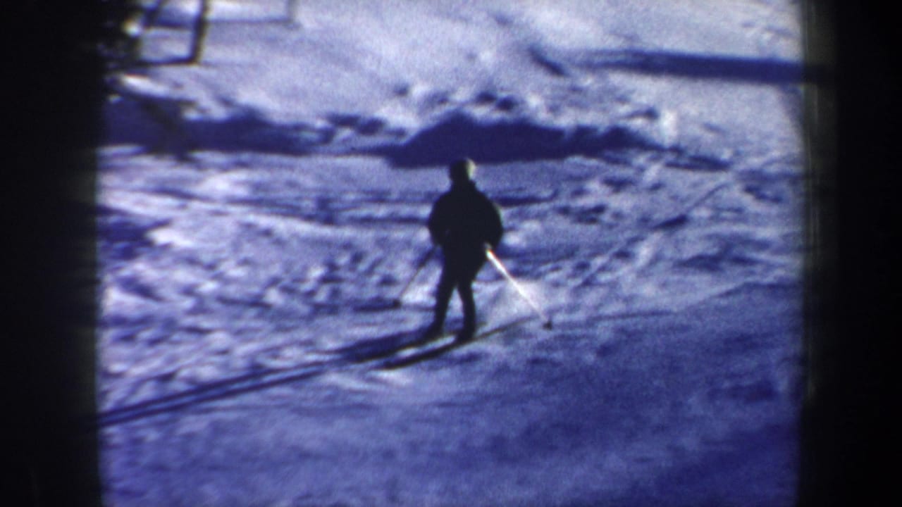The skier is seen closer on the snowy ground, with skis and a long shadow visible in the 8mm frame.