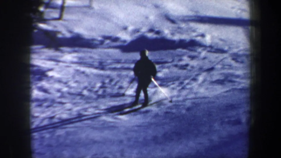The skier is seen closer on the snowy ground, with skis and a long shadow visible in the 8mm frame.