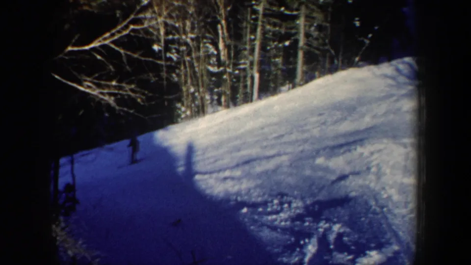 1962 New Hampshire home-movie frame with a lone skier in shadow below a bright snowy forest slope.