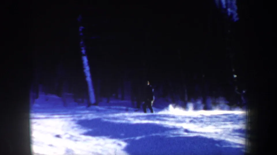 1962 New Hampshire 8mm frame showing a distant skier in dark clothing crossing snowy ground among tall winter trees.