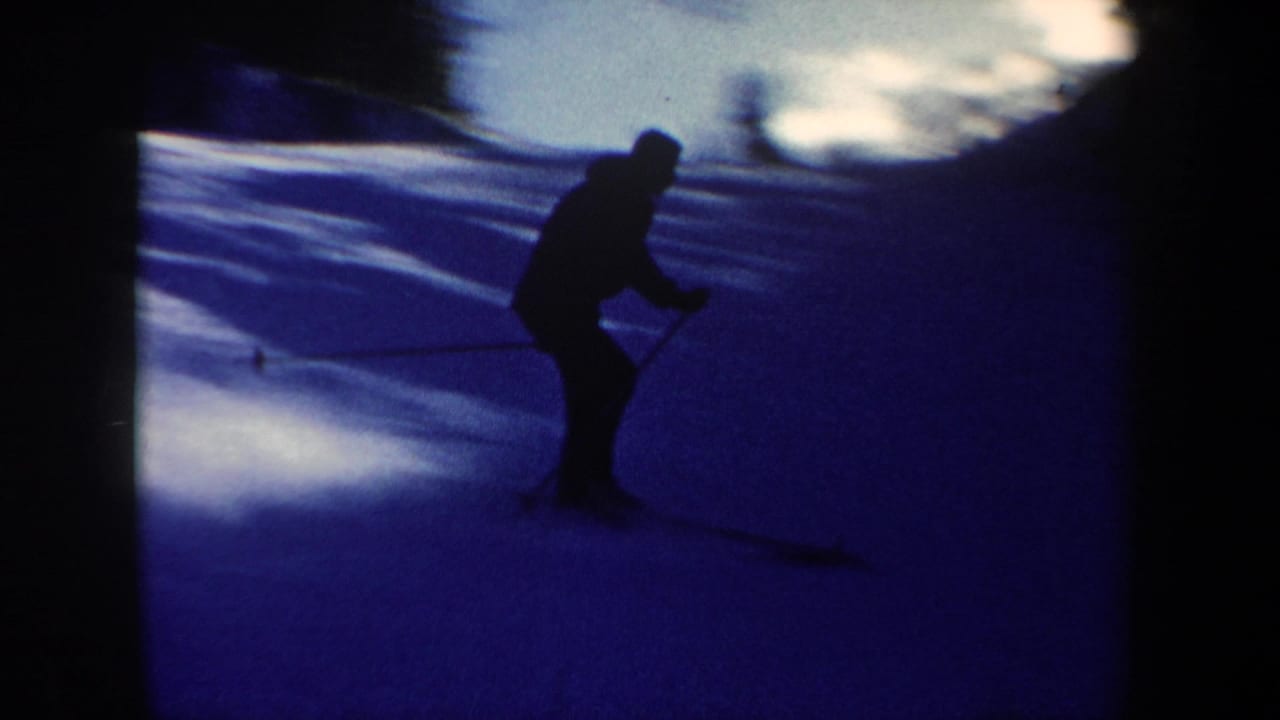 Closer 8mm view of the skier in dark winter clothing using poles while moving across a shadowed snow slope.
