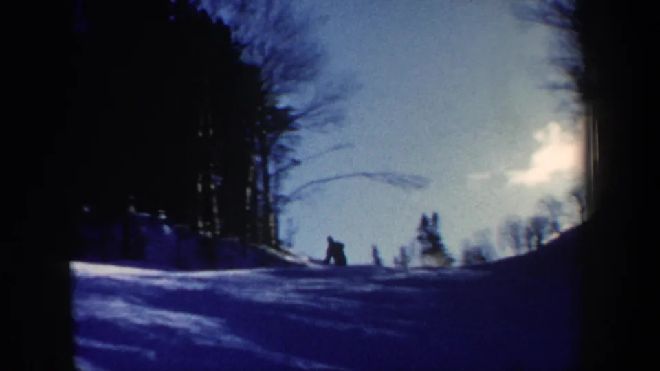 Wide 1962 New Hampshire home-movie frame with a small skier figure on a snowy hillside below bare trees and bright sky.