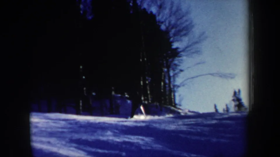 1962 New Hampshire 8mm frame showing a snowy forest slope with a small distant skier and bare trees against a blue winter sky.
