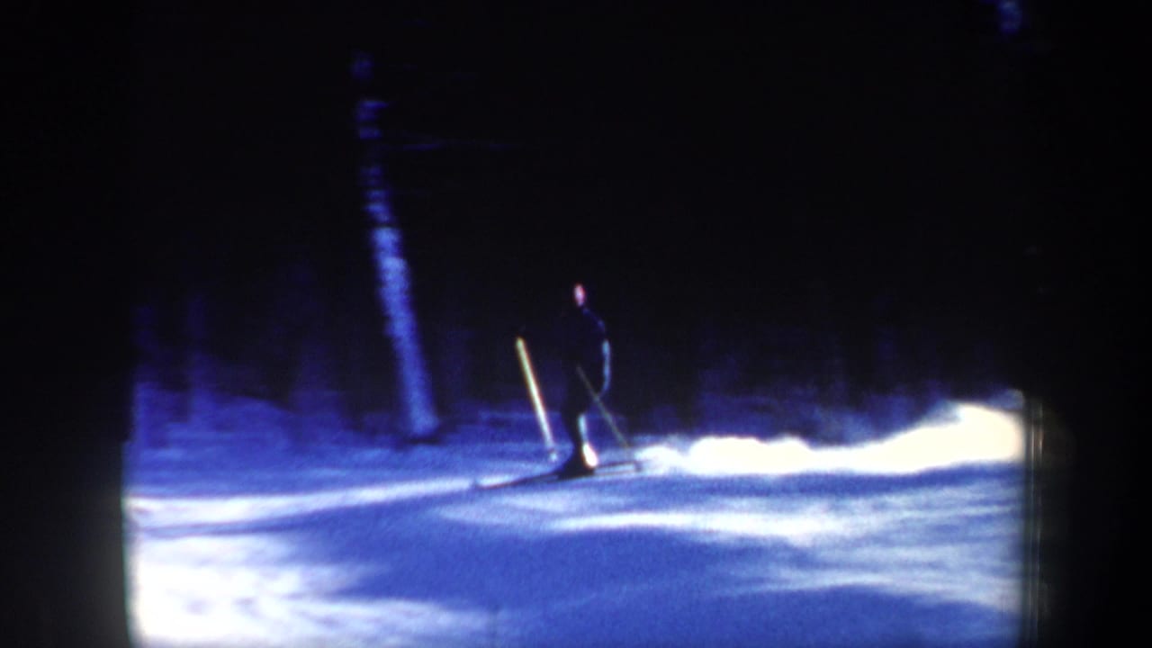Skier in dark clothing gliding across a bright snowy slope beside a dark forest.