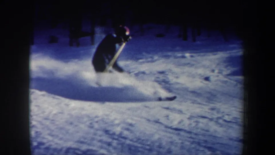 1962 New Hampshire 8mm frame showing a skier in dark winter clothing and red hat carving through powder on a snowy slope.