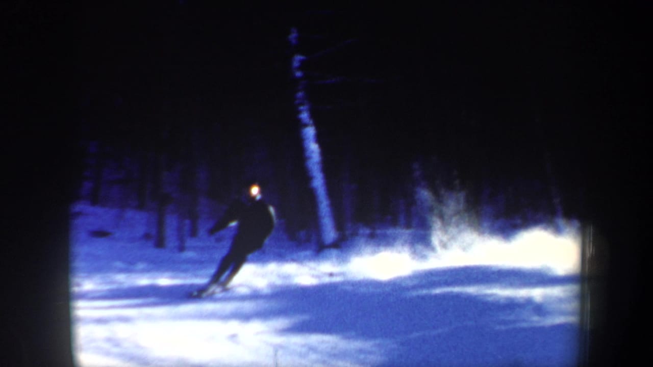 A skier leans through a turn on a snowy slope, with sprayed snow and dark winter woods visible in the 1962 home-movie frame.