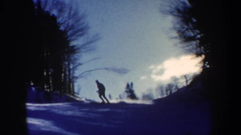 A lone skier descends a snowy New Hampshire hillside in silhouette, with bare trees and a bright clouded sky behind them.