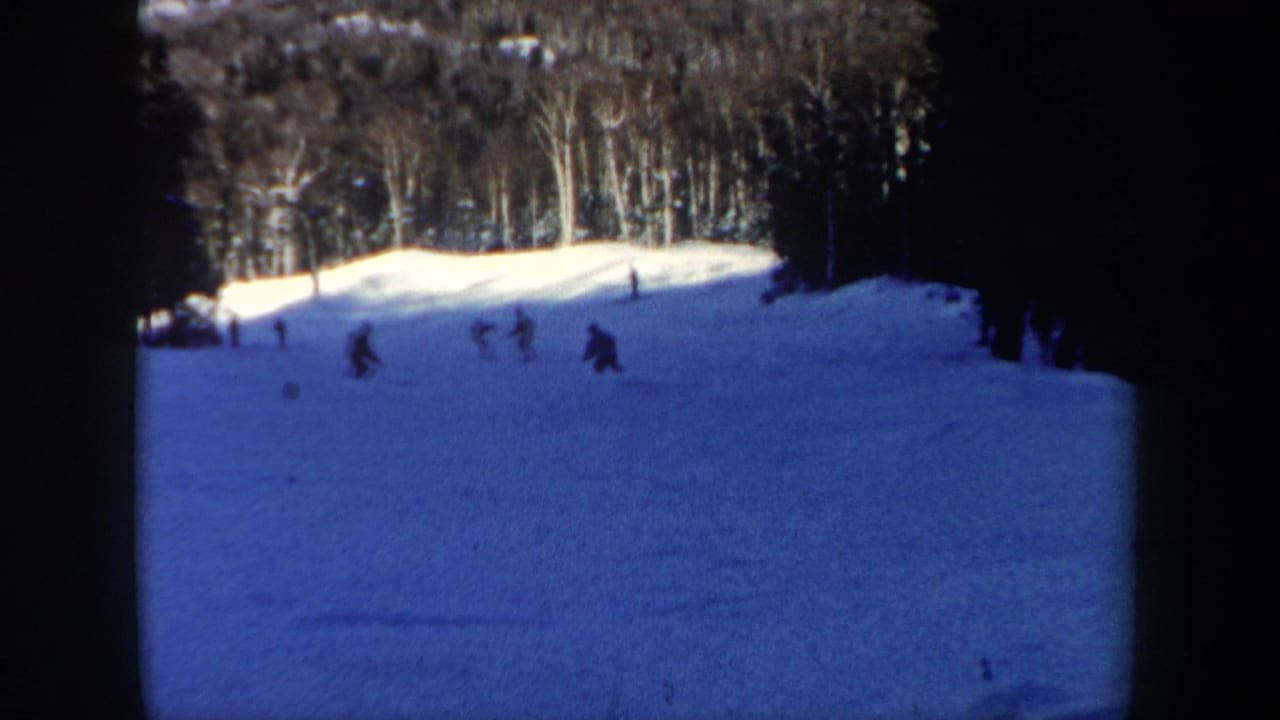 1962 New Hampshire 8mm frame showing small figures skiing down a snow-covered slope with bare winter trees beyond.