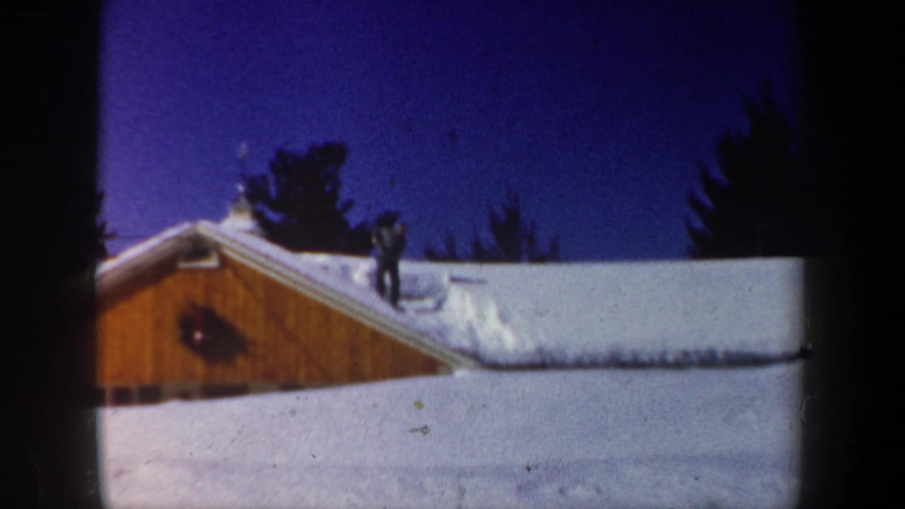1962 New Hampshire 8mm frame showing a person on a snowy wooden building roof, likely clearing snow under a deep blue sky.